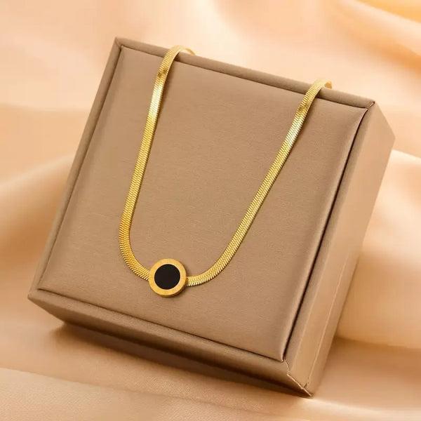 cushioned jewelry box set on a silky beige fabric. In the center of the necklace is a single, circular charm with a deep black inlay and a polished gold border, lying flat to display its minimalist design.