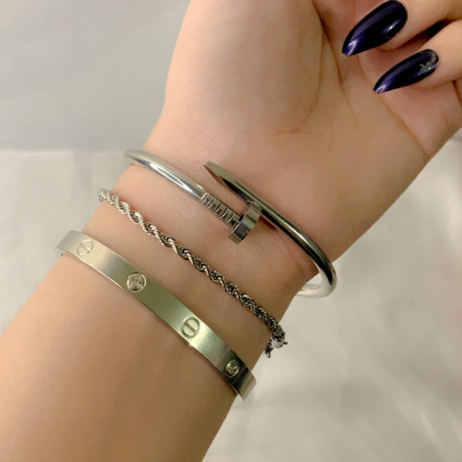 A close-up view of three stylish stainless steel bracelets stacked together, including a silver cuff, a twist chain bracelet, and a bangle with circular accents. The nails are painted in a deep purple shade, adding a sophisticated touch to the ensemble. The bracelets are waterproof and designed for everyday wear.