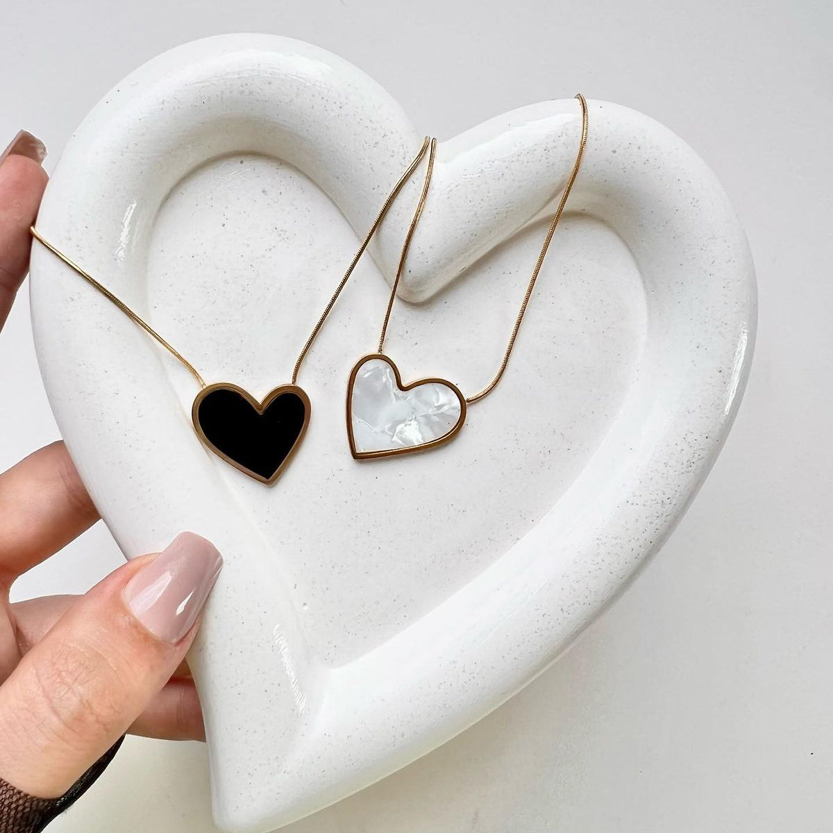 Two gold-framed heart pendant necklaces, one black and one pearl white, displayed on a white ceramic heart tray; waterproof and anti-tarnish premium jewelry set.