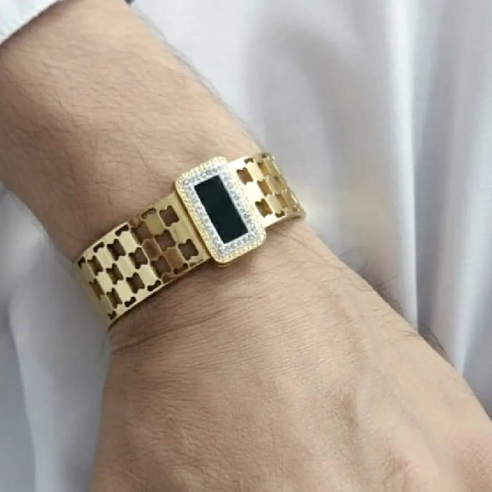 A man's wrist wearing a wide, gold-tone bracelet. The bracelet features a unique checkered cutout link pattern and a large, rectangular centerpiece with a black inlay, bordered by sparkling clear crystals.