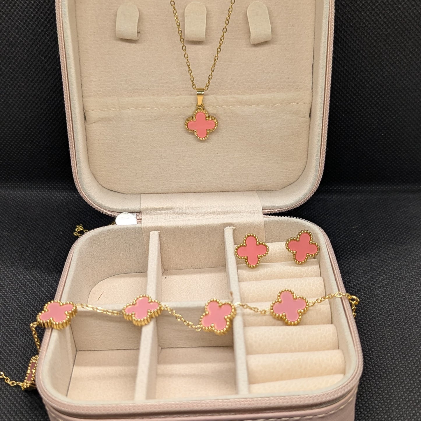 A 3-piece set of gold-toned jewelry featuring a pink enamel four-petal (clover) motif, including a necklace, a charm bracelet, and stud earrings. The set is displayed in an open pink travel jewelry box, emphasizing the waterproof and 70% off sale.