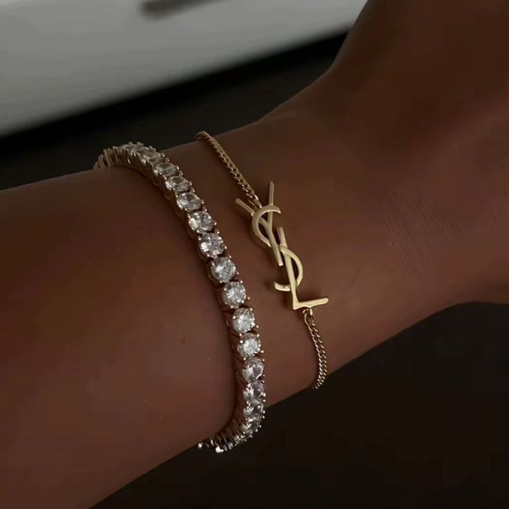 Two stacked gold-tone bracelets: a classic, sparkling crystal tennis bracelet and a fine chain bracelet with a designer-inspired, interlocking initials 'YSL' monogram charm.