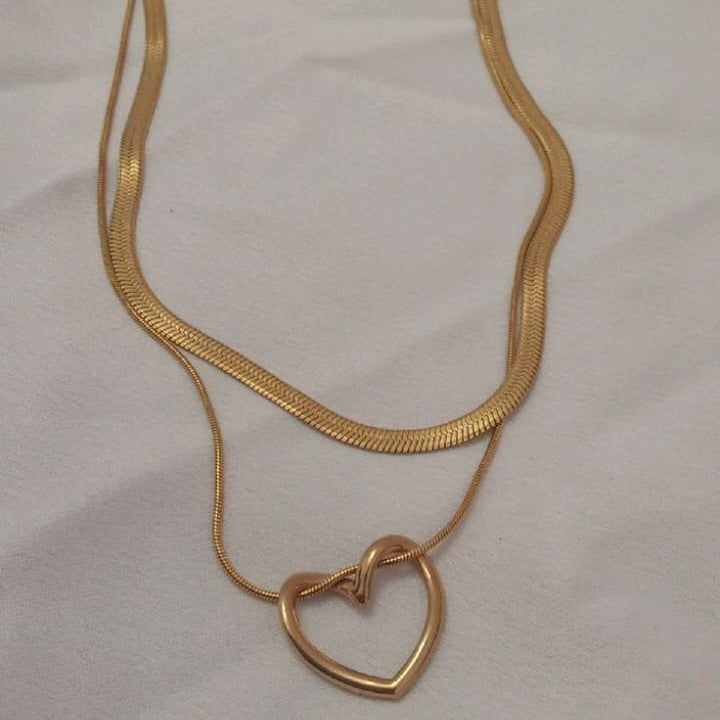 The necklace features two chains joined at the clasp: a shorter, wider, flat herringbone (snake) chain, and a longer, thinner round chain. The longer chain holds a modern, sculptural, 3D open-heart pendant.