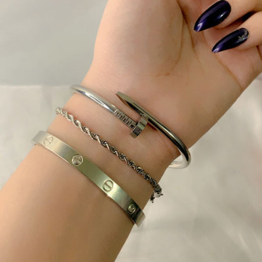A close-up view of three stylish stainless steel bracelets stacked together, including a silver cuff, a twist chain bracelet, and a bangle with circular accents. The nails are painted in a deep purple shade, adding a sophisticated touch to the ensemble. The bracelets are waterproof and designed for everyday wear.