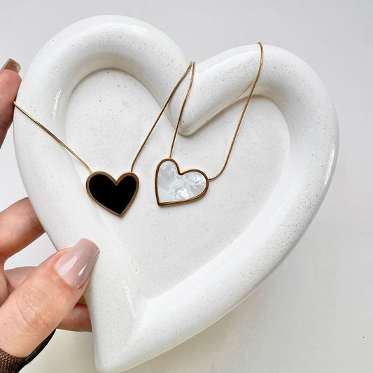 Two gold-framed heart pendant necklaces, one black and one pearl white, displayed on a white ceramic heart tray; waterproof and anti-tarnish premium jewelry set.
