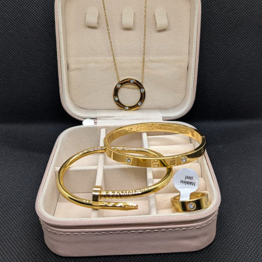 A 4-piece set of gold-toned jewelry made of Stainless Steel, including a circle pendant necklace, two iconic-style bangles (one with clear stones and one in a nail/clou design), and a stone-accented ring. The set is displayed in an open pink travel jewelry box, emphasizing the waterproof and 70% off sale