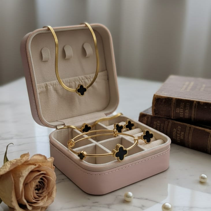 A 5-piece gold-toned jewelry set featuring a black enamel four-petal (clover) motif, including a snake chain necklace, a bangle, a charm bracelet, and stud earrings. The set is displayed in an open pink travel jewelry box on a marble surface, highlighting the waterproof and 70% off sale.