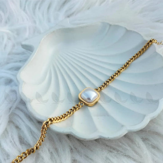 Gold-plated chain bracelet with a square white pearl centerpiece displayed on a white seashell dish over a soft, fluffy background.
