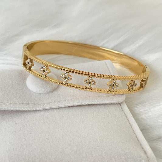 The cuff features a detailed openwork design with four repeated quatrefoil floral clusters, each intricately set with small, sparkling pave cubic zirconia stones. The bracelet is resting on a soft, light grey velvet jewelry pouch on a fluffy white background.