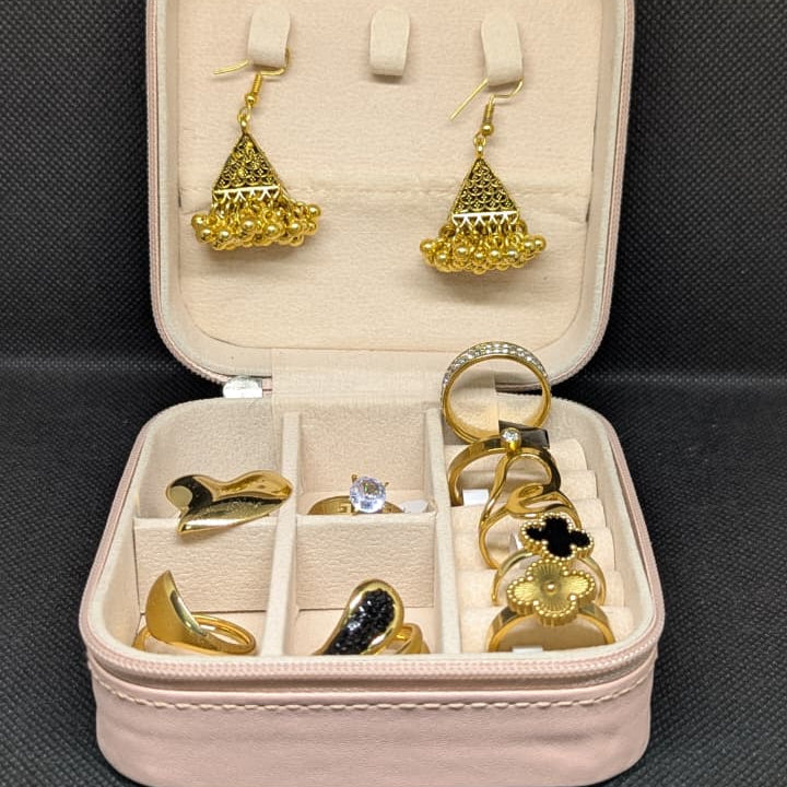 A large 9-piece gold-toned jewelry collection displayed in a pink travel box. The set includes three pairs of earrings (puffed heart, square black enamel, and gold beaded jhumka) and six rings in various styles (pave stone, solitaire, black clover, and gold bands). The set is 100% waterproof (metal items) and offered at 70% off.