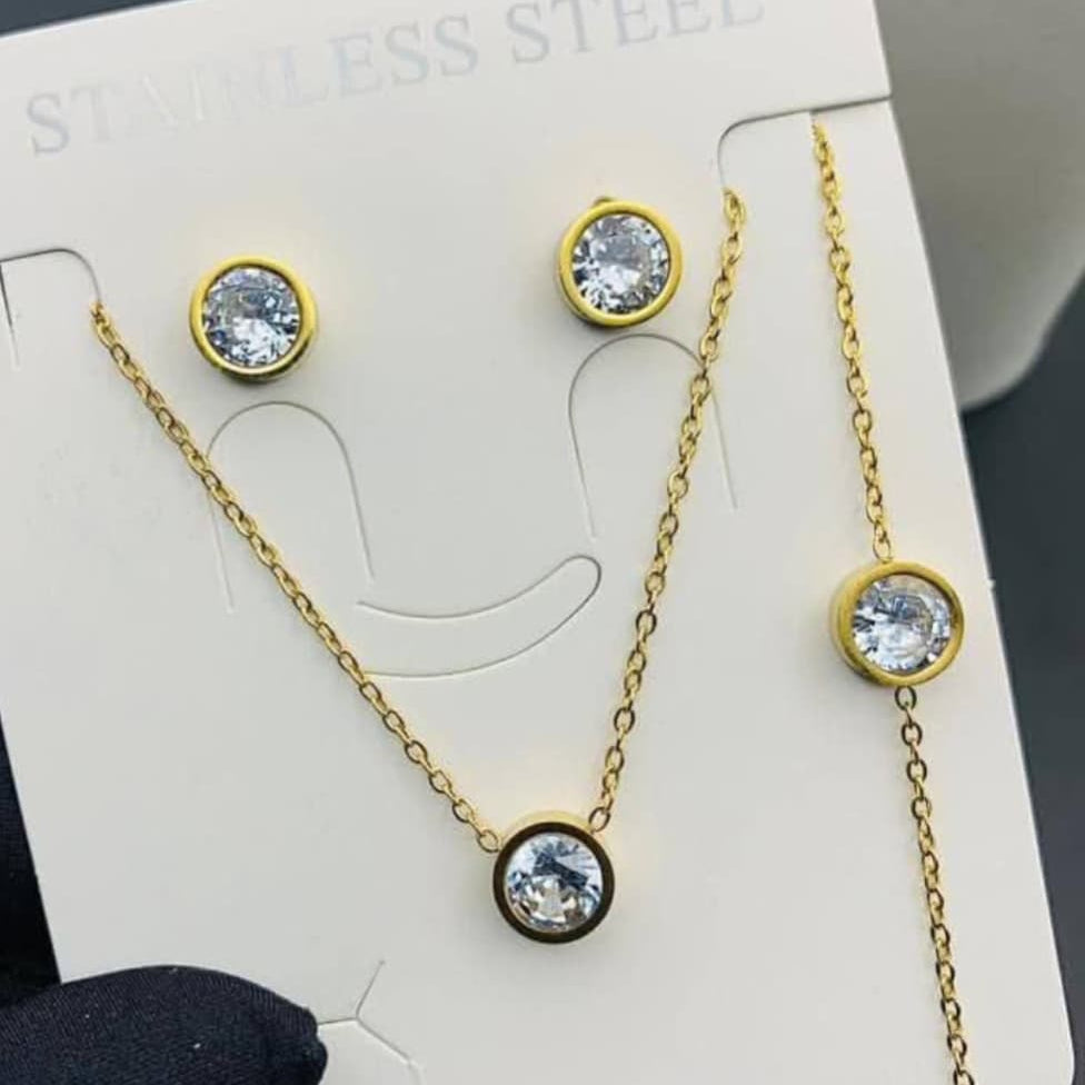The set includes a gold-tone necklace, a matching chain bracelet, and a pair of stud earrings. Each piece features a single, round, brilliant-cut cubic zirconia stone set in a sleek gold bezel setting.
