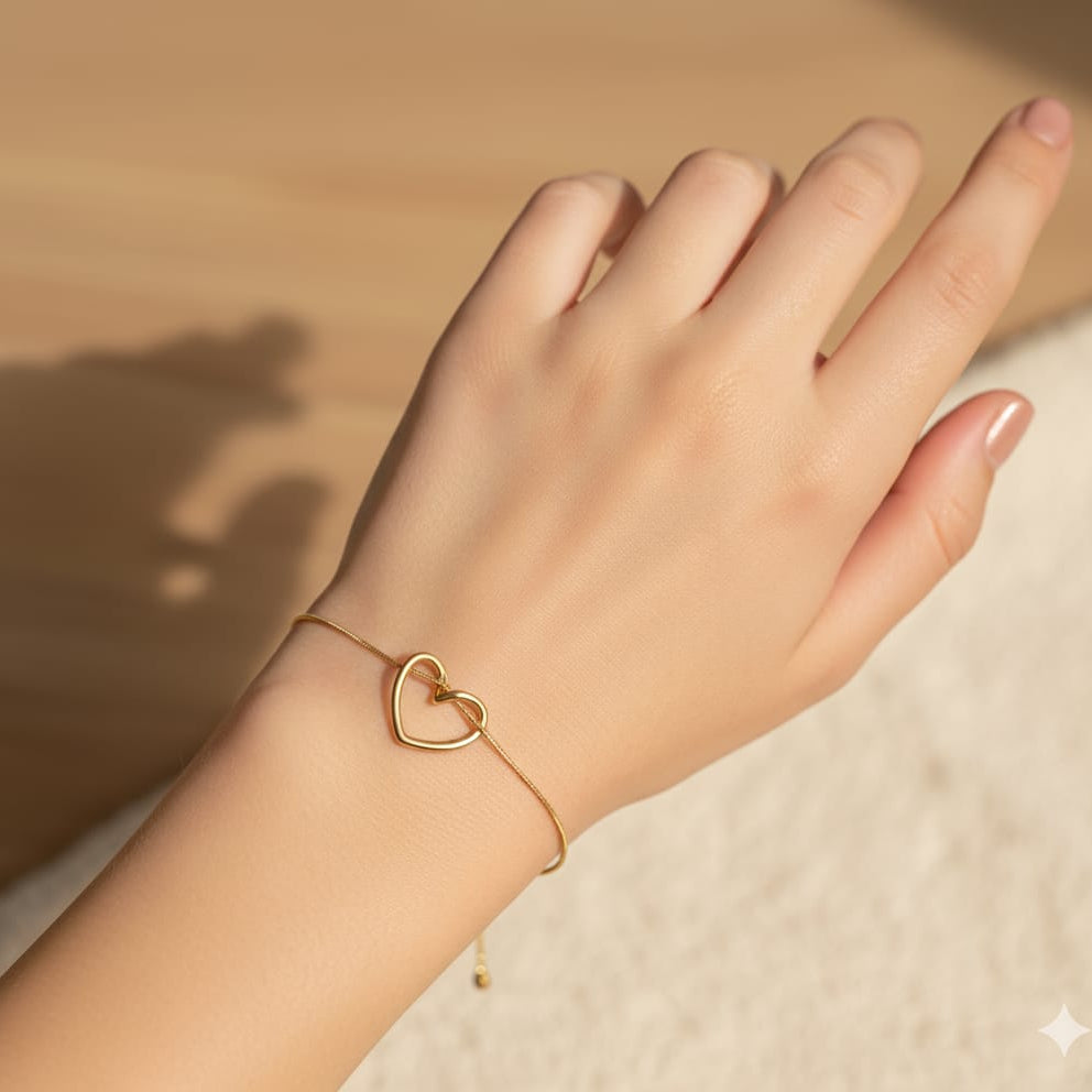 Close-up shot of a delicate gold chain bracelet featuring a small, minimalist open-outline heart charm worn on a fair wrist under natural sunlight.