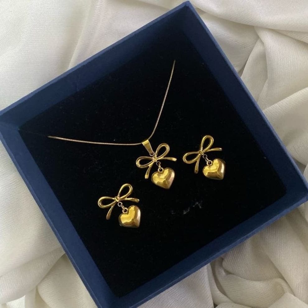 A gold jewelry set featuring a bow and heart pendant necklace with matching bow-and-heart earrings, displayed in a blue gift box on soft white fabric; waterproof and anti-tarnish premium jewelry.