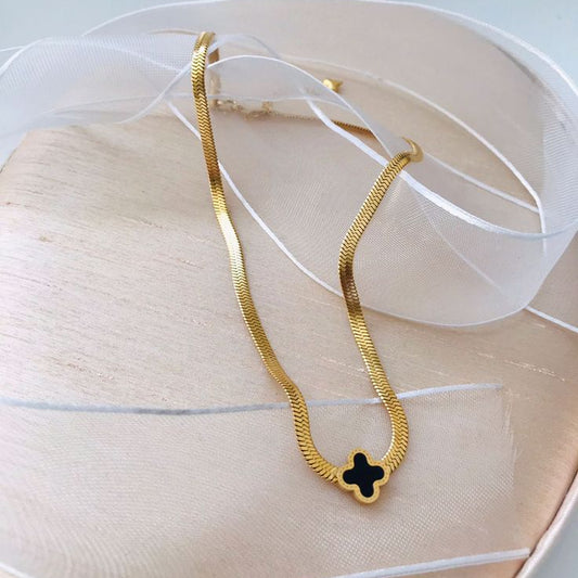 A high-angle shot of a gold-tone, flat snake chain (herringbone) necklace, resting on a beige satin cushion. In the center of the chain is a small, four-leaf clover (quatrefoil) charm, which has a black inlay and a polished gold border. A sheer white ribbon is draped artfully in the background, adding a soft, elegant touch.