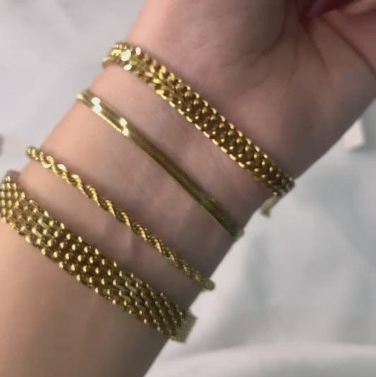 A hand showcasing a collection of five elegant gold bracelets: a delicate beaded gold chain, a smooth wire bangle, a twisted gold bangle, a ball-chain bracelet, and a classic gold link chain. The bracelets are waterproof, anti-tarnish, and designed for everyday wear.